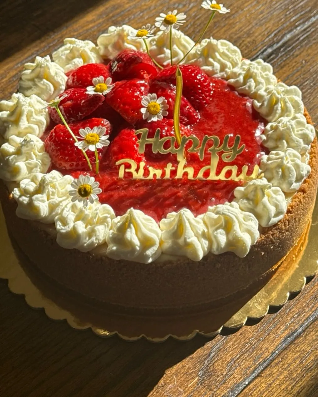 A strawberry cheesecake decorated with a gold 'Happy Birthday' topper, fresh strawberries and daisies.